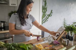 Personne préparant un repas à domicile, coupant des légumes sur une planche tout en suivant une recette sur une tablette.