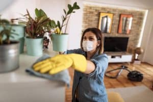 Personne masquée nettoyant une étagère dans un salon moderne, avec des plantes et un aspirateur en arrière-plan.