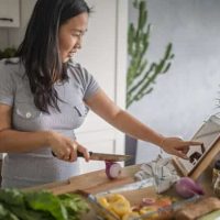 Personne préparant un repas à domicile, coupant des légumes sur une planche tout en suivant une recette sur une tablette.