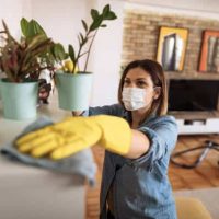 Personne masquée nettoyant une étagère dans un salon moderne, avec des plantes et un aspirateur en arrière-plan.